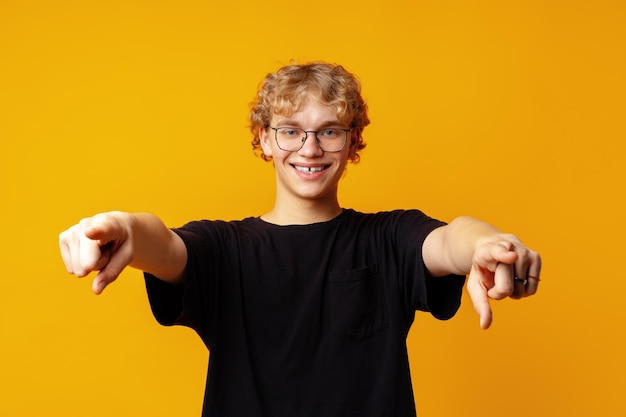 Young curlyhaired man in glasses pointing to camera against yellow background