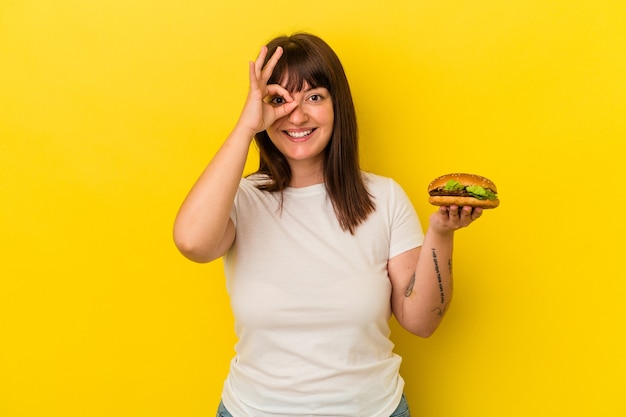 Молодая соблазнительная кавказская женщина, держащая гамбургер, изолирована на желтом фоне, взволнована, держа на глазах одобренный жест.