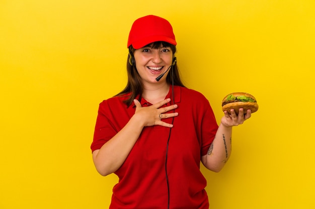Молодая пышная кавказская женщина, держащая гамбургер на желтом фоне, громко смеется, держа руку на груди.