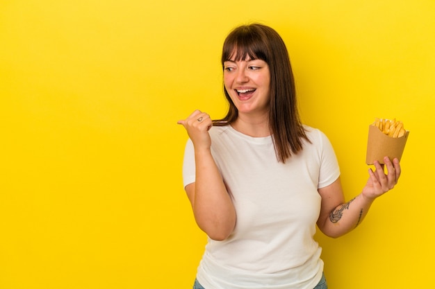 Young curvy caucasian woman holding fries isolated on yellow background points with thumb finger away, laughing and carefree