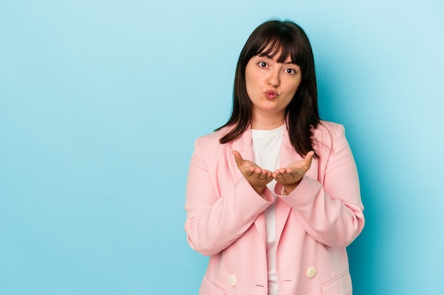 Young curvy caucasian woman isolated on blue background folding lips and holding palms to send air kiss.