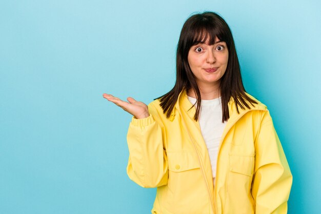 Young curvy caucasian woman isolated on blue background showing a copy space on a palm and holding another hand on waist.