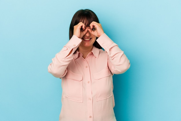 Young curvy caucasian woman isolated on blue background showing okay sign over eyes