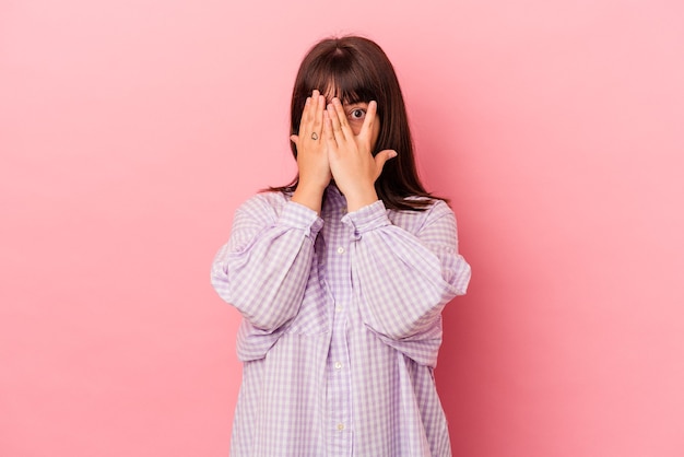 Young curvy caucasian woman isolated on pink background blink through fingers frightened and nervous.