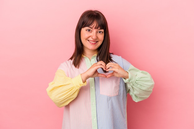 Young curvy caucasian woman isolated on pink background smiling and showing a heart shape with hands.