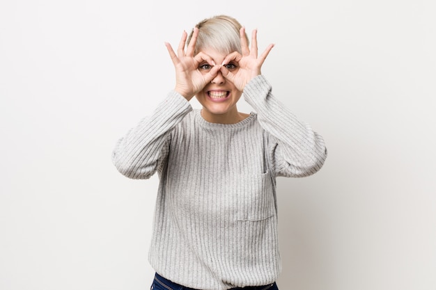 Young curvy caucasian woman isolated on white wall showing okay sign over eyes