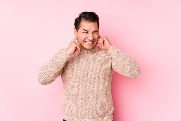 Young curvy man posing in a pink wall isolated covering ears with hands.