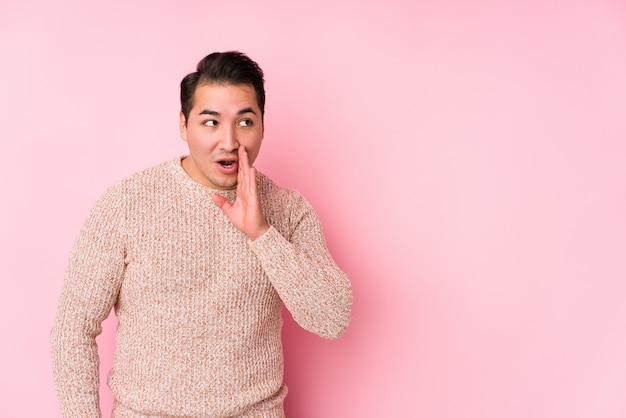 Young curvy man posing in a pink wall isolated is saying a secret hot braking news and looking aside