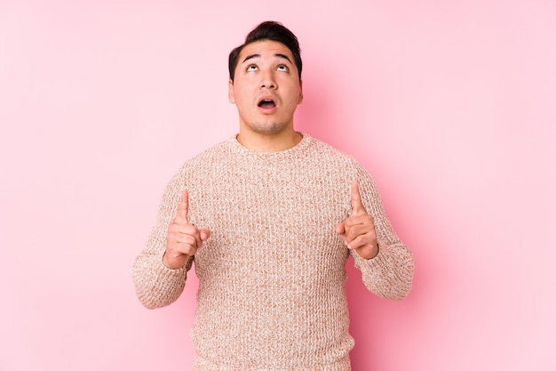 Young curvy man posing in a pink wall isolated pointing upside with opened mouth.