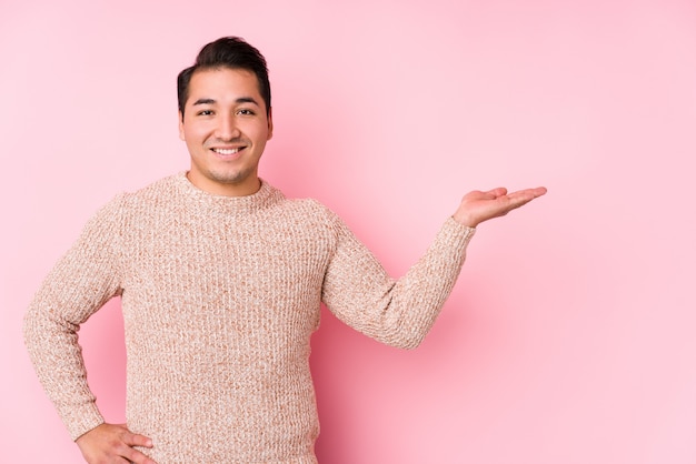 Young curvy man posing in a pink wall isolated showing a copy space on a palm and holding another hand on waist.