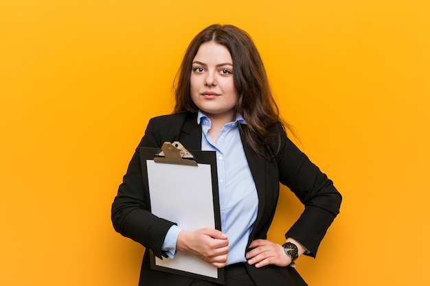 Young curvy plus size business woman holding a clipboard smiling confident with crossed arms.