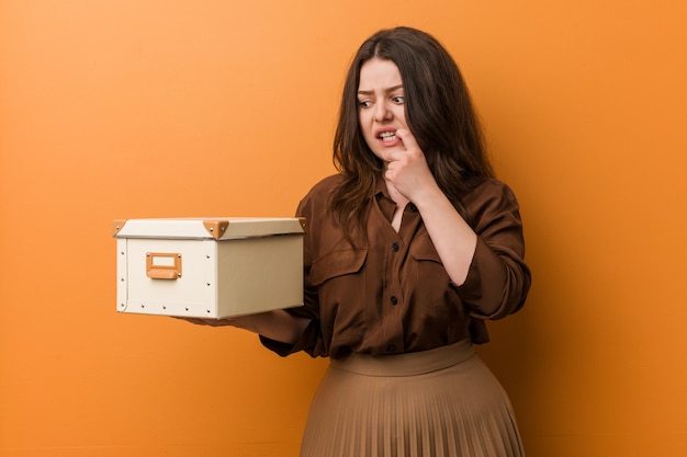 Young curvy plus size woman holding a box biting fingernails, nervous and very anxious.