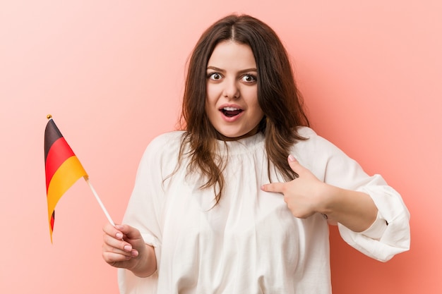 Young curvy plus size woman holding a germany flag surprised pointing at himself, smiling broadly.