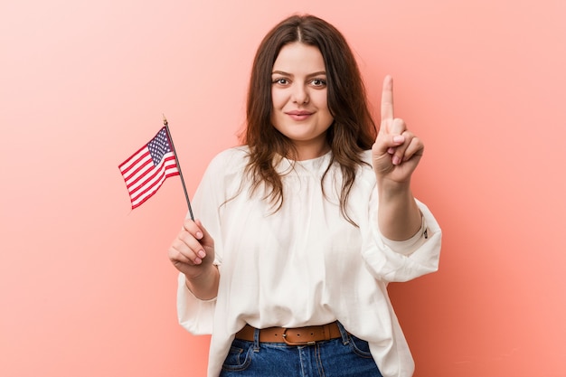 Young curvy plus size woman holding a united states flag showing number one with finger