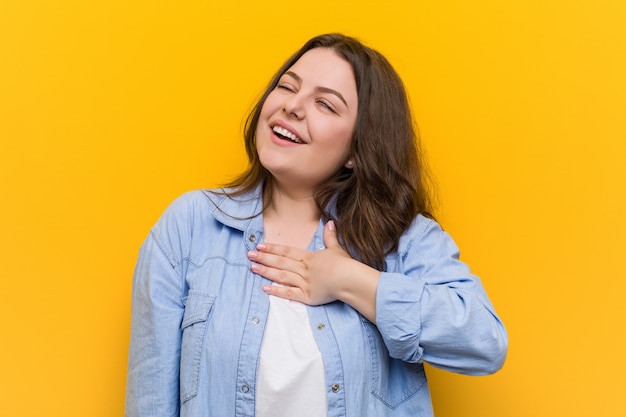 Young curvy plus size woman laughs out loudly keeping hand on chest.