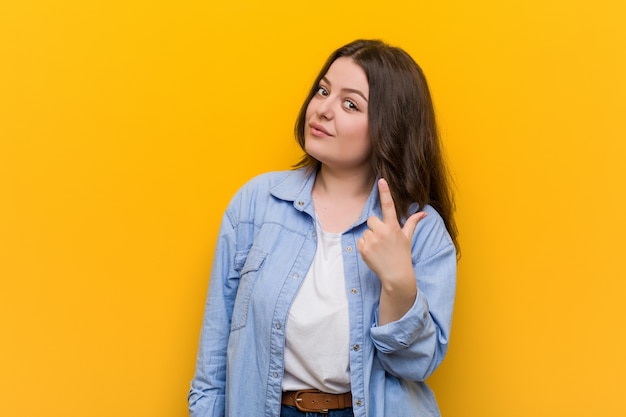 Young curvy plus size woman pointing with finger at you as if inviting come closer.