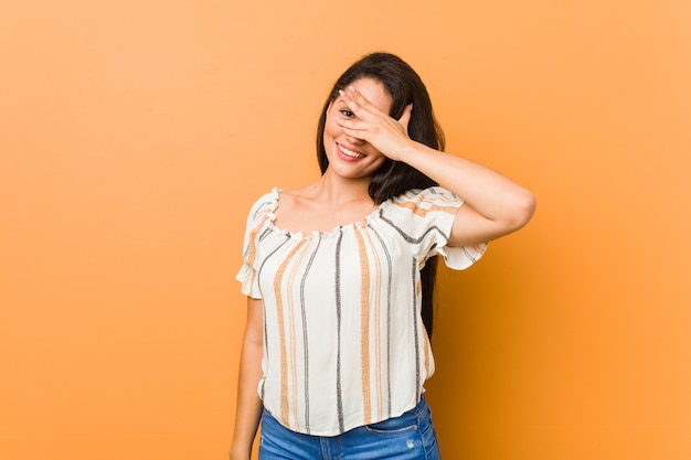 Young curvy woman blink at the camera through fingers, embarrassed covering face.