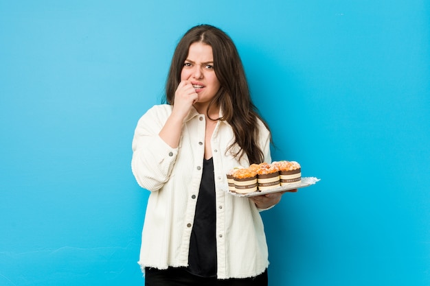 Young curvy woman holding a cupcakes biting fingernails, nervous and very anxious.