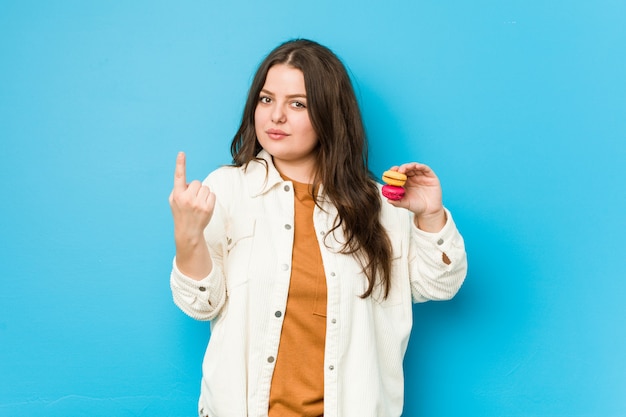 Young curvy woman holding macaroons pointing with finger at you as if inviting come closer.