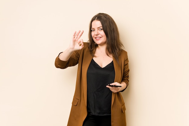 Young curvy woman holding a phone cheerful and confident showing ok gesture