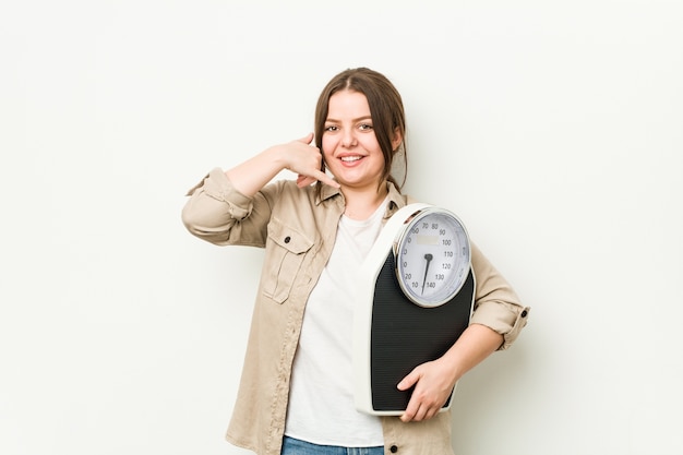 Young curvy woman holding a scale showing a mobile phone call gesture with fingers.