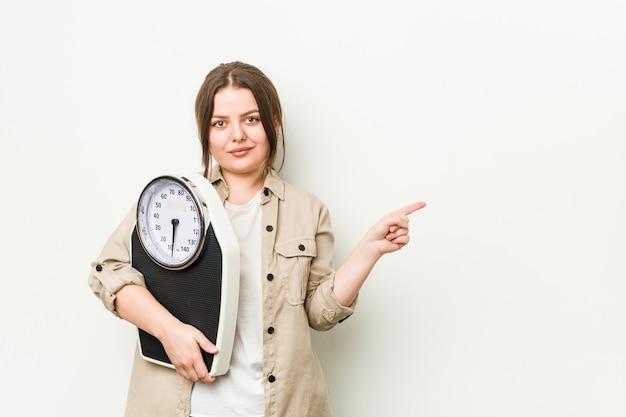 Young curvy woman holding a scale smiling and pointing aside, showing something at blank space.