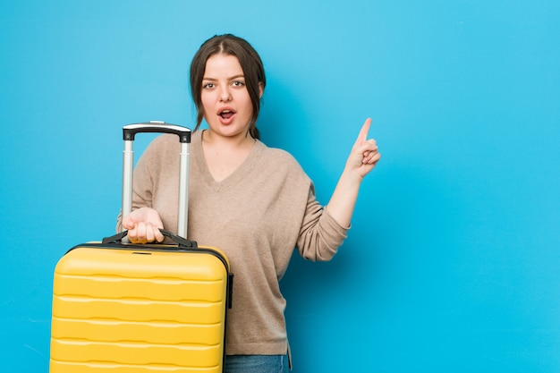 Young curvy woman holding a suitcase pointing to the side