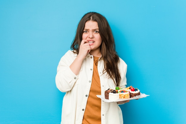 Young curvy woman holding a sweet cakes biting fingernails, nervous and very anxious.