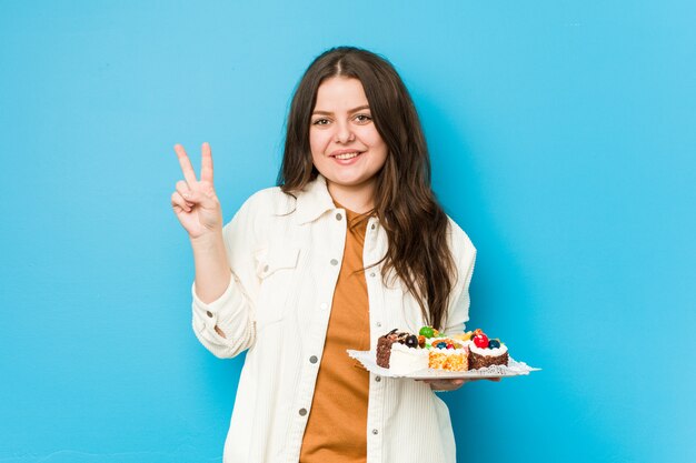 Young curvy woman holding a sweet cakes showing number two with fingers.