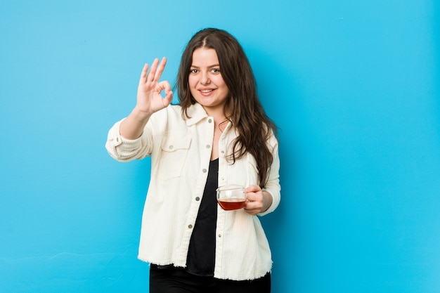 Young curvy woman holding a tea cup cheerful and confident showing ok gesture.