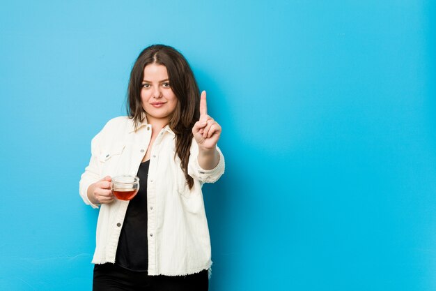 Young curvy woman holding a tea cup showing number one with finger.