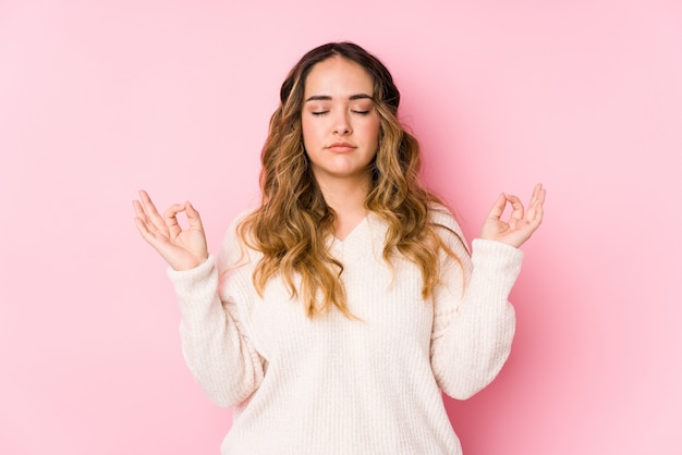 Young curvy woman posing in a pink wall isolated relaxes after hard working day, she is performing yoga.