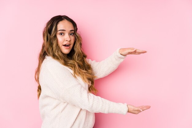 Foto giovane donna curvy che posa in una parete rosa isolata colpita e stupita tenendo uno spazio della copia fra le mani.