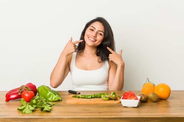 Young curvy woman preparing a healthy meal smiles, pointing fingers at mouth.