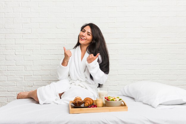 Young curvy woman taking a breakfast on the bed raising both thumbs up, smiling and confident.