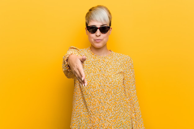 Young curvy woman wearing a floral summer clothes stretching hand at camera in greeting gesture.