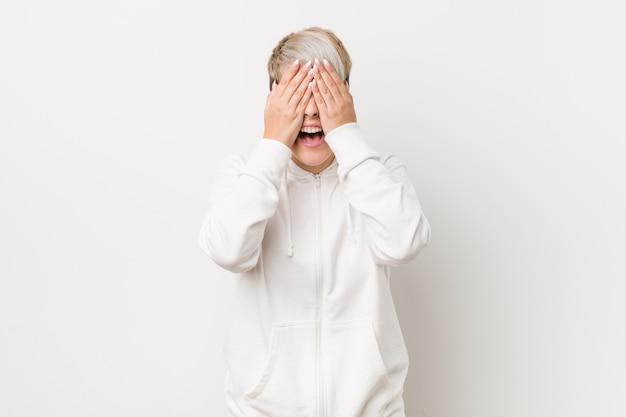 Young curvy woman wearing a white hoodie covers eyes with hands, smiles broadly waiting for a surprise.