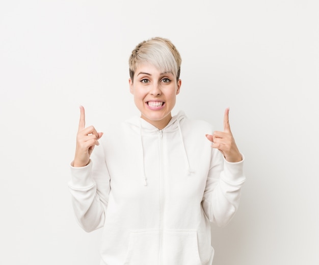 Young curvy woman wearing a white hoodie indicates with both fore fingers up showing a blank space.