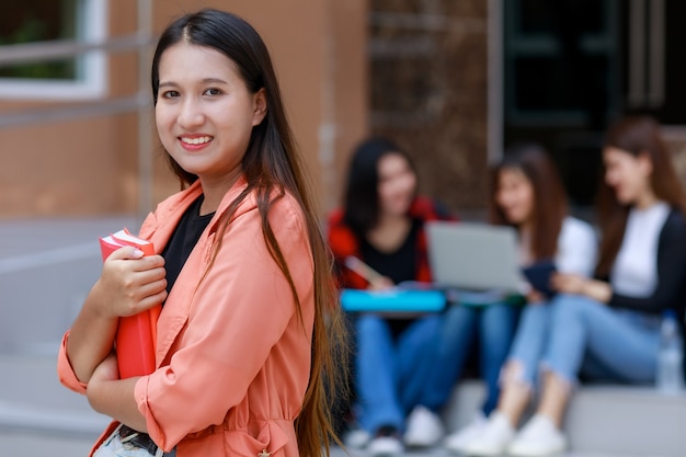 本を持っている若くてかわいいアジアの大学生の女の子は、学校の建物の前で背景にぼやけている友人のグループと一緒にカメラにポーズをとる。十代の若者たちの親友の概念の学習と友情。