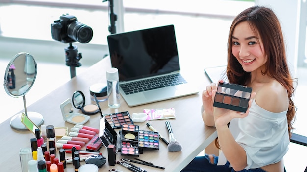 Young and cute Asian female vlogger, influencer, or online seller sitting with cosmetics products and DSLR camera and laptop notebook computer ready to broadcast online live stream or recording video.