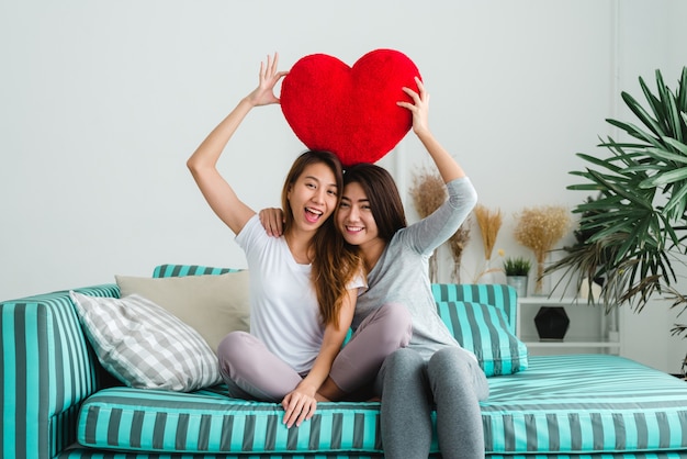Молодые симпатичные азиатские лесбиянки, держащие красную сердечную форму ивы вместе, улыбаясь с счастью у себя дома