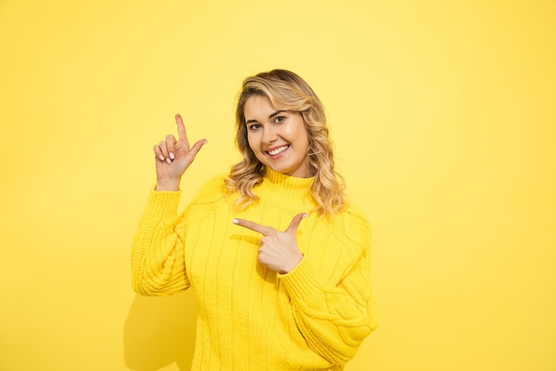 Young cute beautiful blonde surprised woman wearing yellow sweater standing over yellow isolated background, pointing with two hands with fingers to copy space for commersial sign. Studio shot.