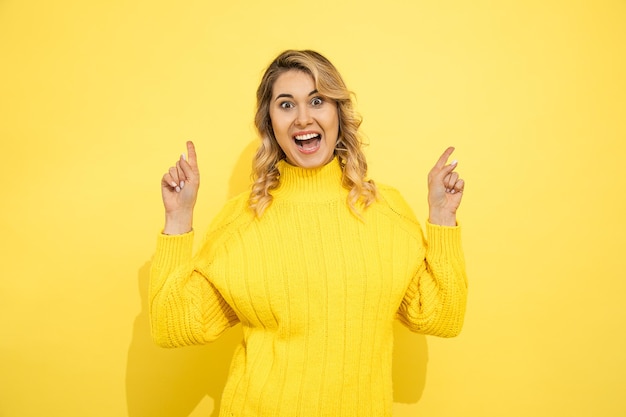 Young cute beautiful happy smiling woman wearing yellow sweater standing over yellow isolated background, pointing with hands to copy space for commersial sign. Positive emocion. Studio shot.