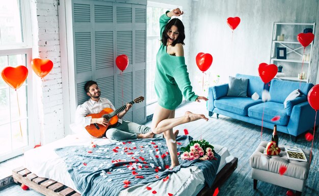 Young cute beautiful woman is dancing on the middle of bedroom while her handsome guy is playing for her on guitar. Love, celebration, romantic, st. Valentine's day, women day, birthday, date