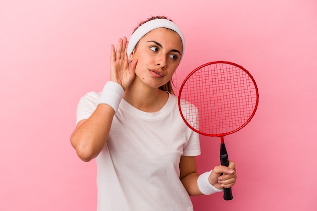 Young cute blonde caucasian woman holding a badminton racket isolated on pink background trying to listening a gossip.