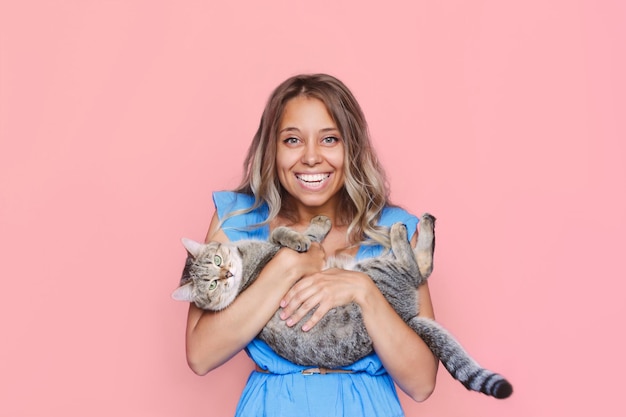 young cute blonde woman holds a cat in her hands like a baby on a light color pink background