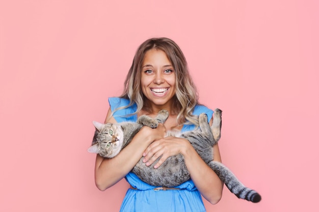 Young cute blonde woman holds a cat in her hands like a baby on a light color pink background