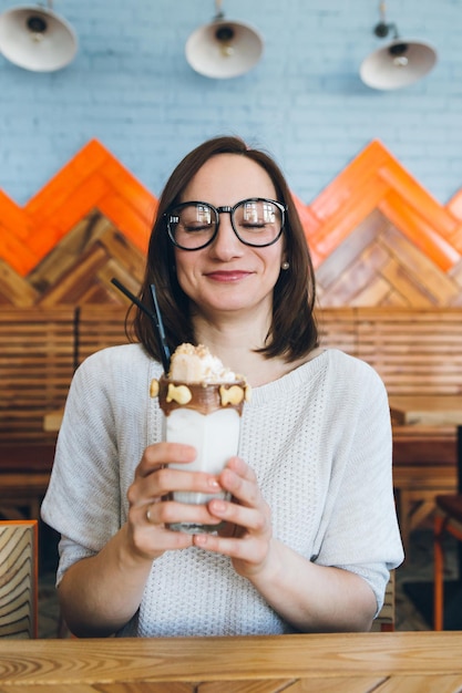 La giovane donna castana sveglia beve il frappè splendidamente decorato con crema e biscotti nella caffetteria. tonificante.