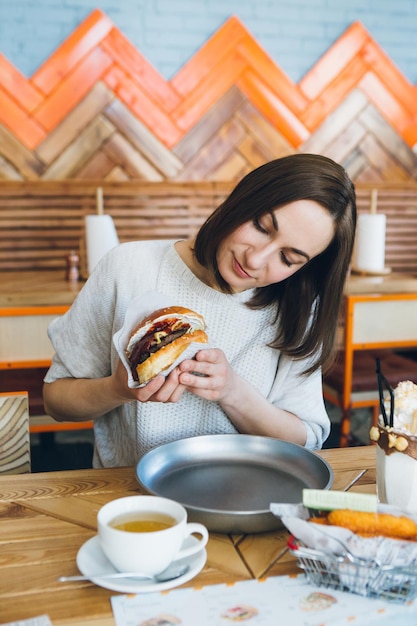 写真 カフェで作りたてのおいしいハンバーガーを食べる若いかわいいブルネットの女性。調色。