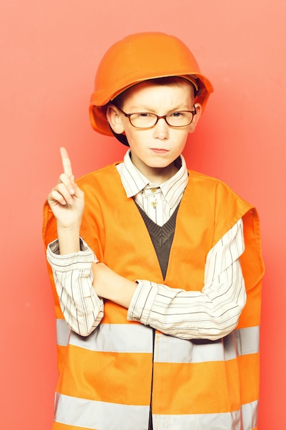 Young cute builder boy in orange uniform and hard hat or helmet with glasses with raised finger on red studio background